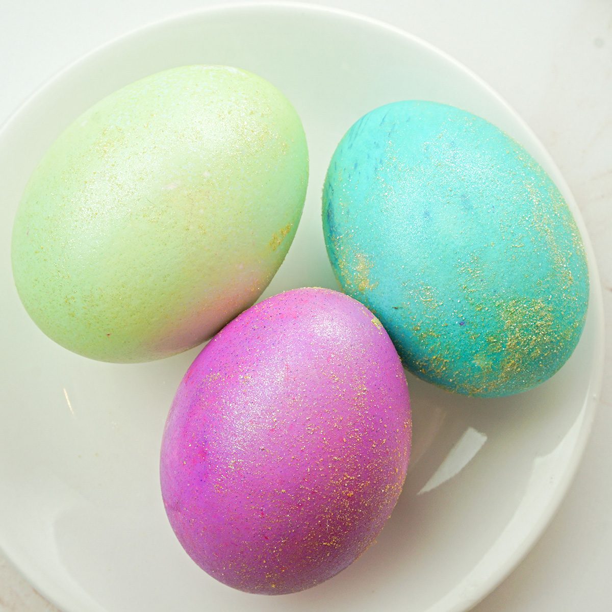 Three pastel-colored eggs, one green, one blue, and one pink, each speckled with a subtle gold pattern, are arranged on a white plate.