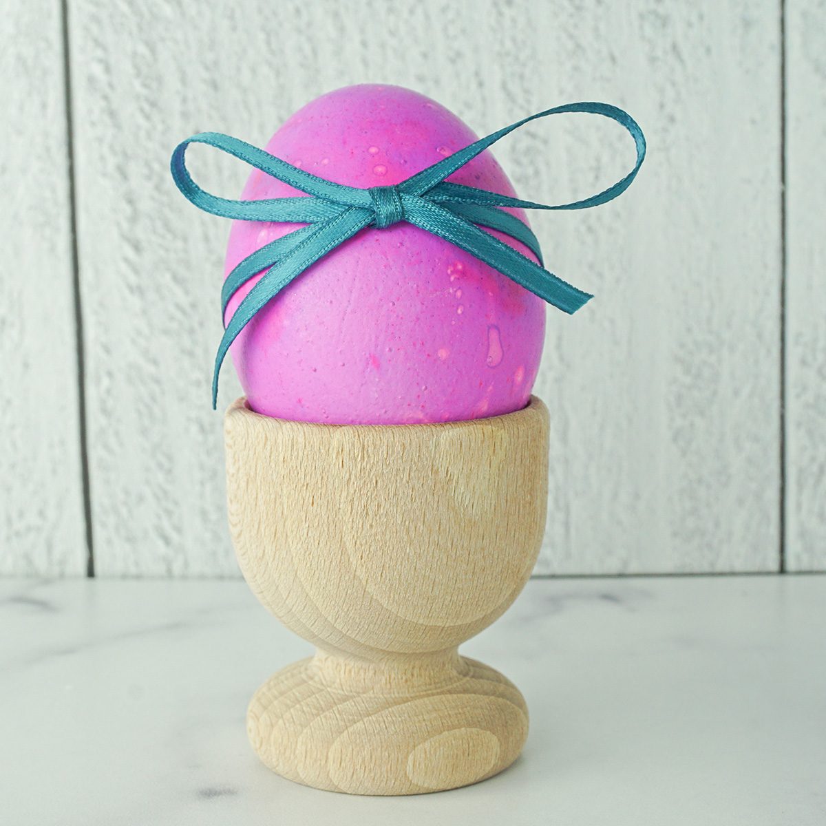 A pink-colored egg with a teal ribbon bow sits in a wooden egg cup. The background features a light wood-textured surface.