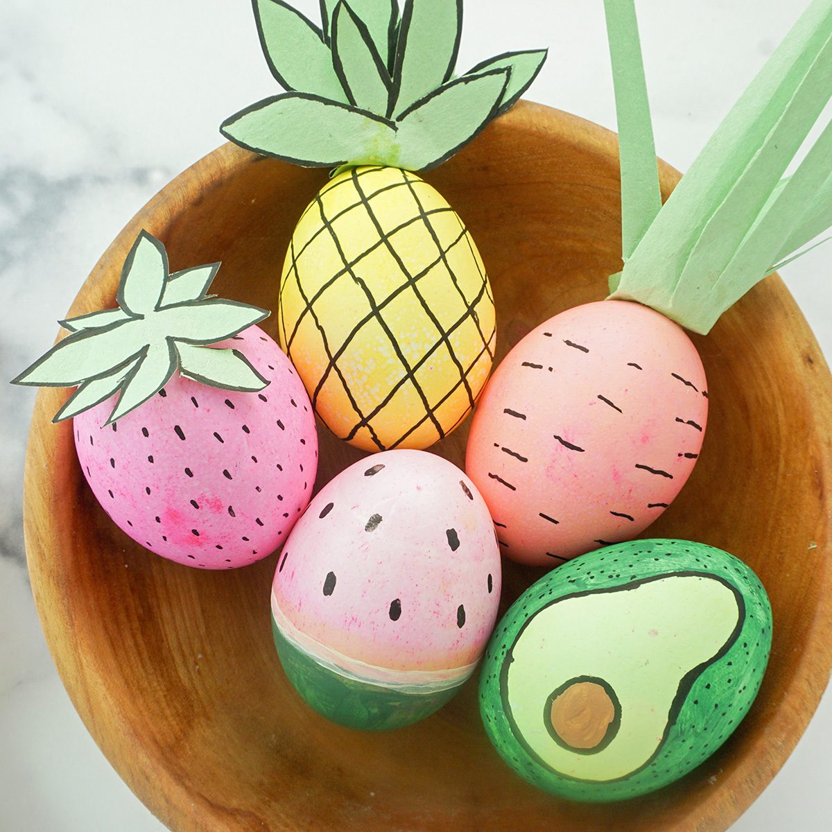 A wooden bowl contains five hand-painted eggs, each decorated as a fruit: a pineapple, strawberry, watermelon, avocado, and an unknown fruit. Each egg has paper leaves attached, enhancing the fruit theme.