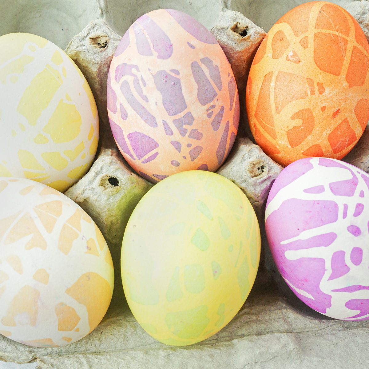 Six colorful, abstractly patterned Easter eggs are arranged in a carton. The eggs feature pastel shades of yellow, pink, orange, and purple with unique designs, creating a festive and artistic appearance.