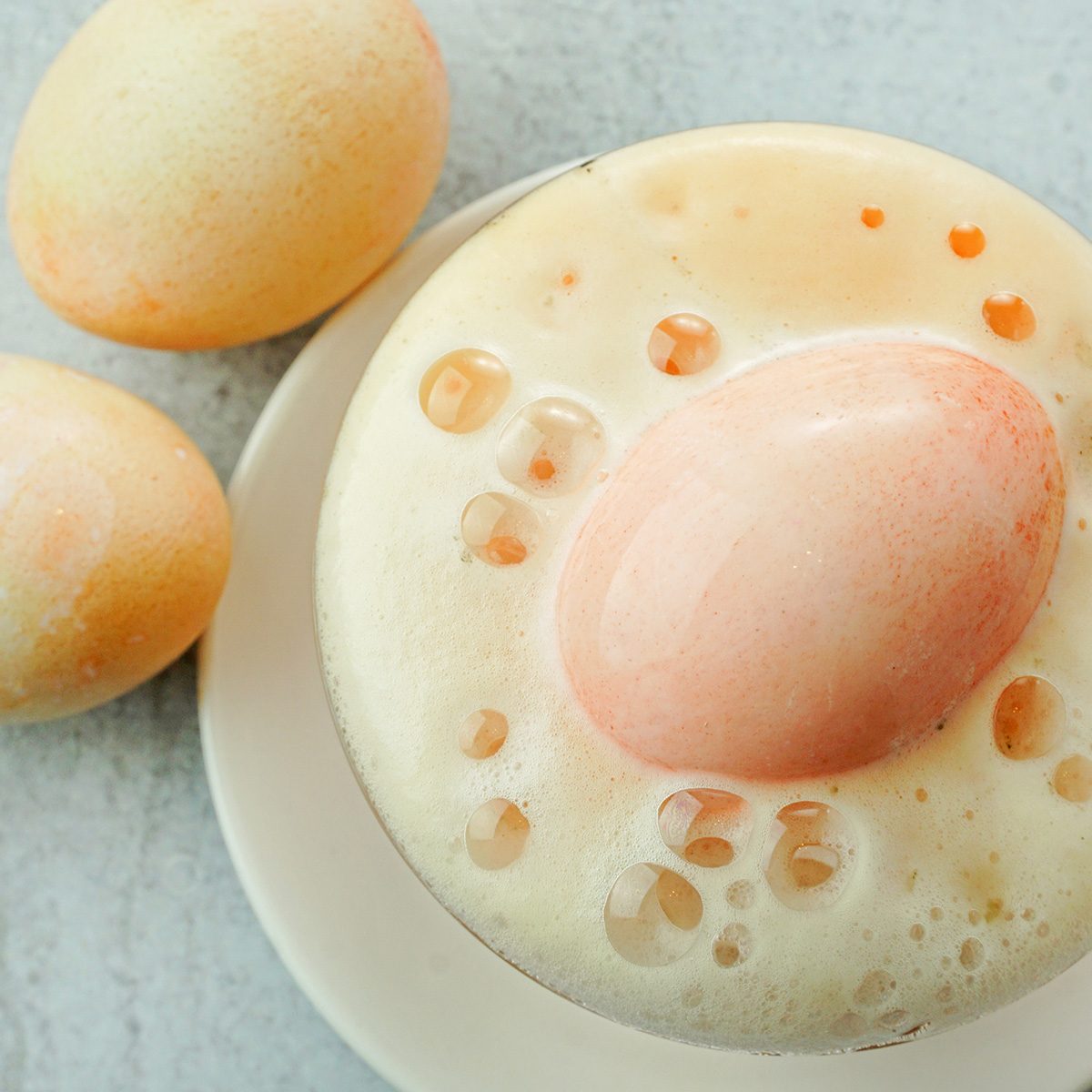 Three eggs are shown, two whole and one partially submerged in a foamy liquid within a white bowl on a light gray surface. The foam has bubbles surrounding the visible egg.