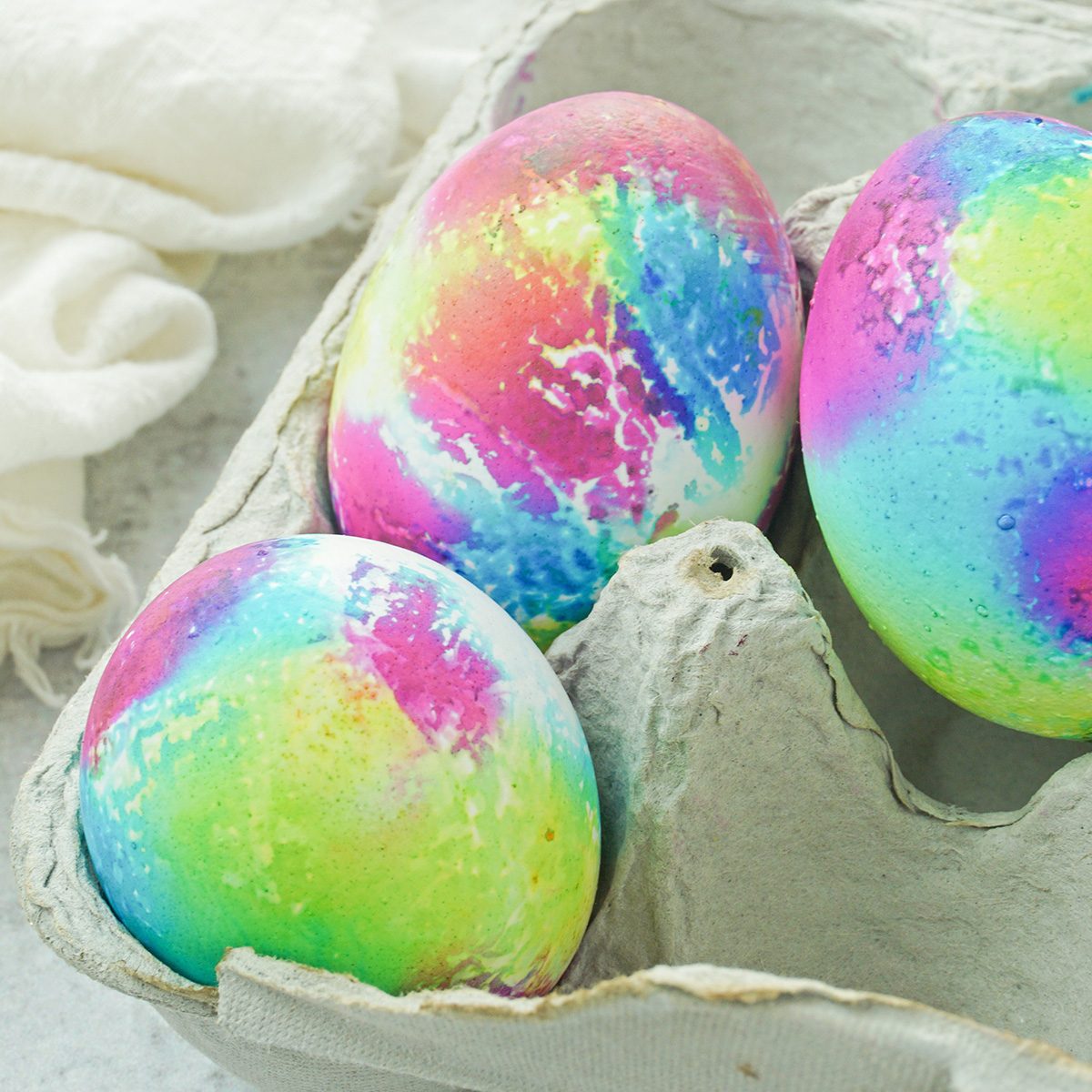 Three eggs in a carton, painted with vibrant, marbled colors including pink, blue, green, and yellow, creating a rainbow effect. A soft white cloth is in the background.
