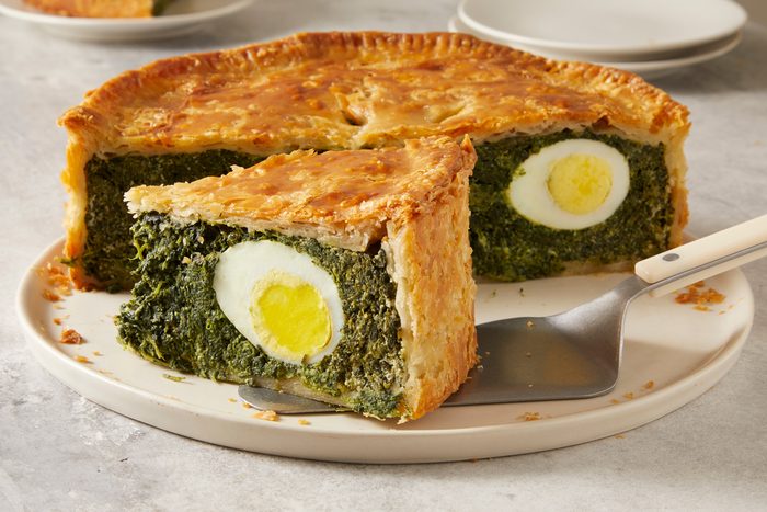 Table view shot of Torta Pasqualina; placed on a large plate; cut into pieces with cake and served on a small plate; empty plates in the background; all set on marble surface;