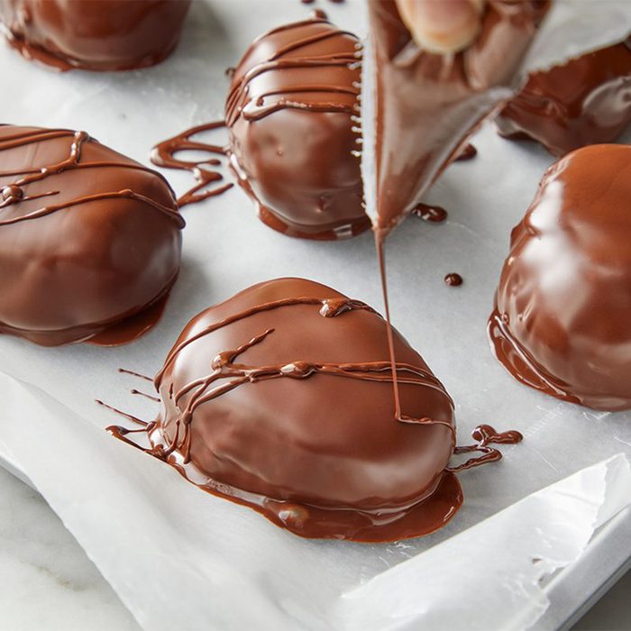 A person drizzles chocolate over several chocolate-covered treats on parchment paper. The treats are oval-shaped, glossy, and placed on a baking tray.
