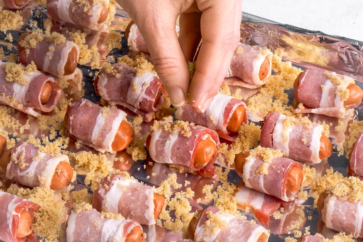 brown sugar being sprinkled over bacon wrapped sausages