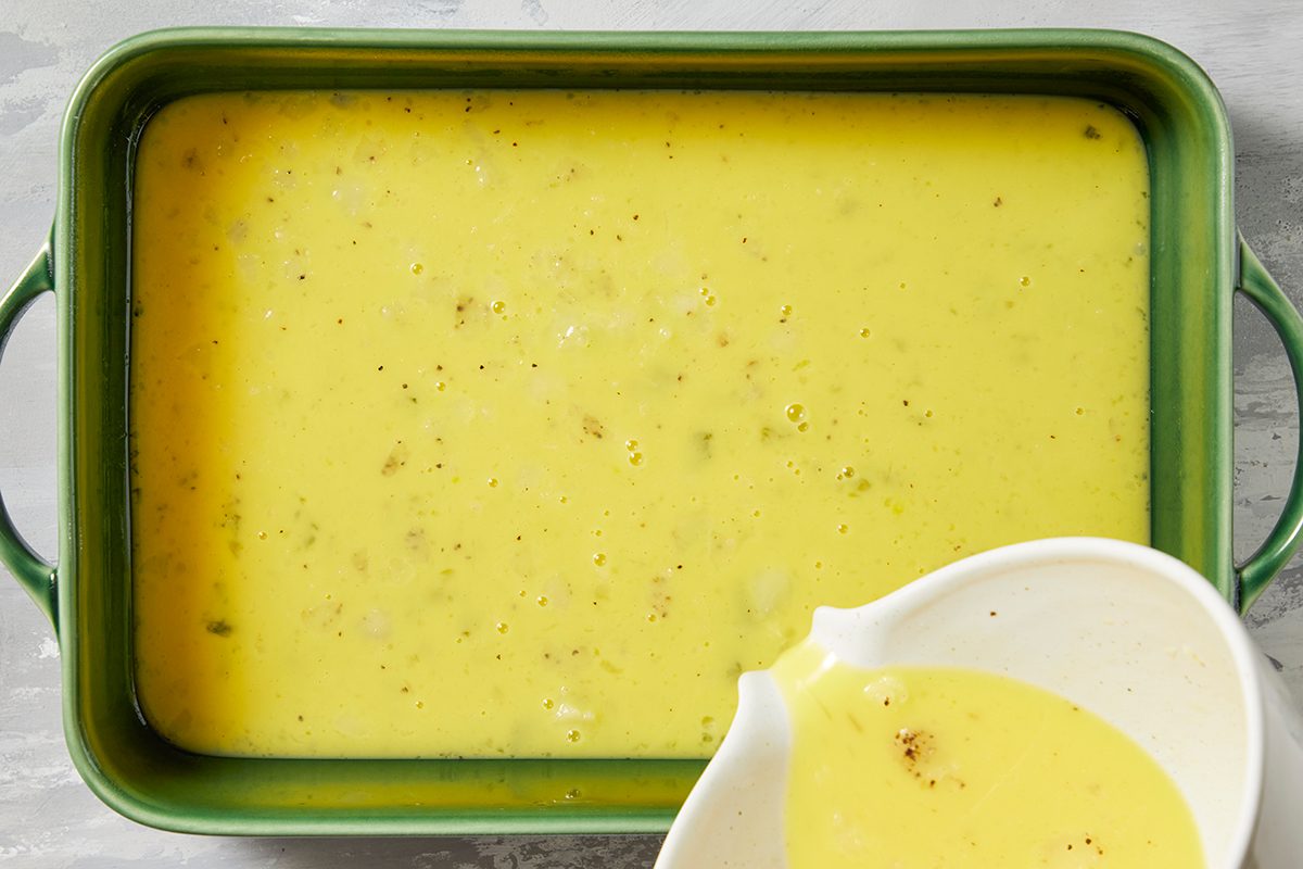 A green rectangular baking dish filled with creamy yellow soup, featuring small specks of seasoning. A white pouring jug, partially visible, contains the same soup. 