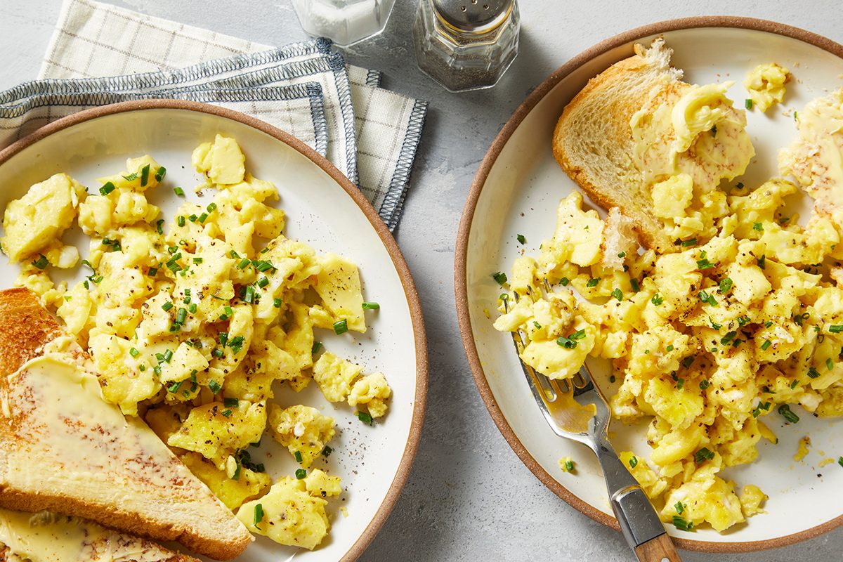 Two plates of scrambled eggs garnished with herbs are on a table. Each plate includes a piece of buttered toast. 