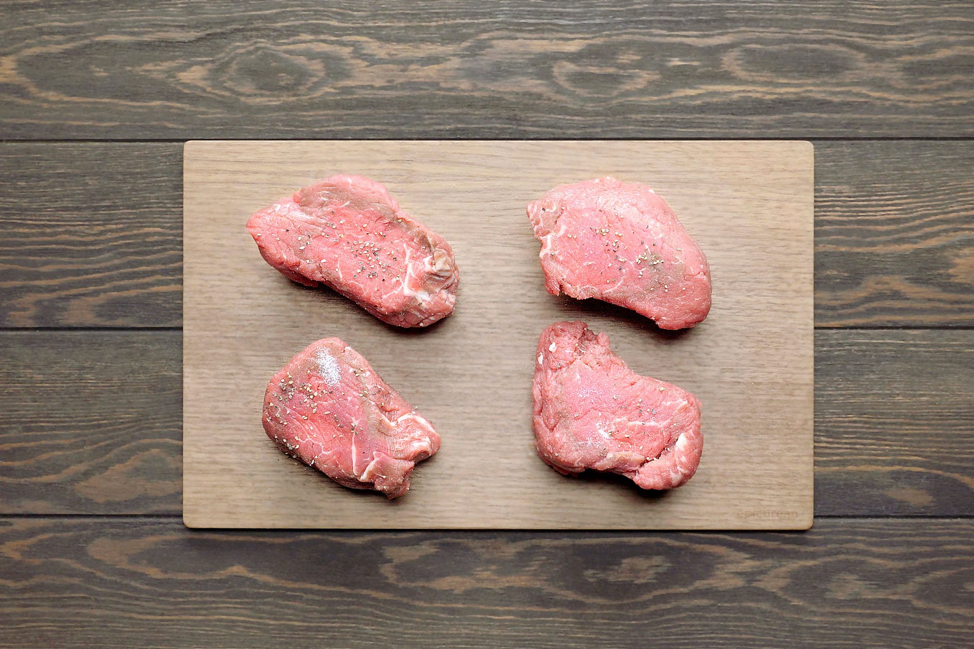 overhead shot of four raw beef filets on a wooden cutting board, the cutting board is on a brown wooden table