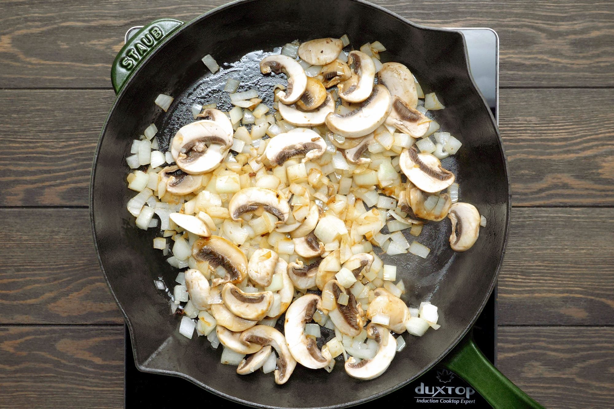 overhead shot of a cast iron skillet with a green handle, sitting on a wooden countertop; the skillet is filled with chopped onions and sliced mushrooms that are being sauteed in oil;
