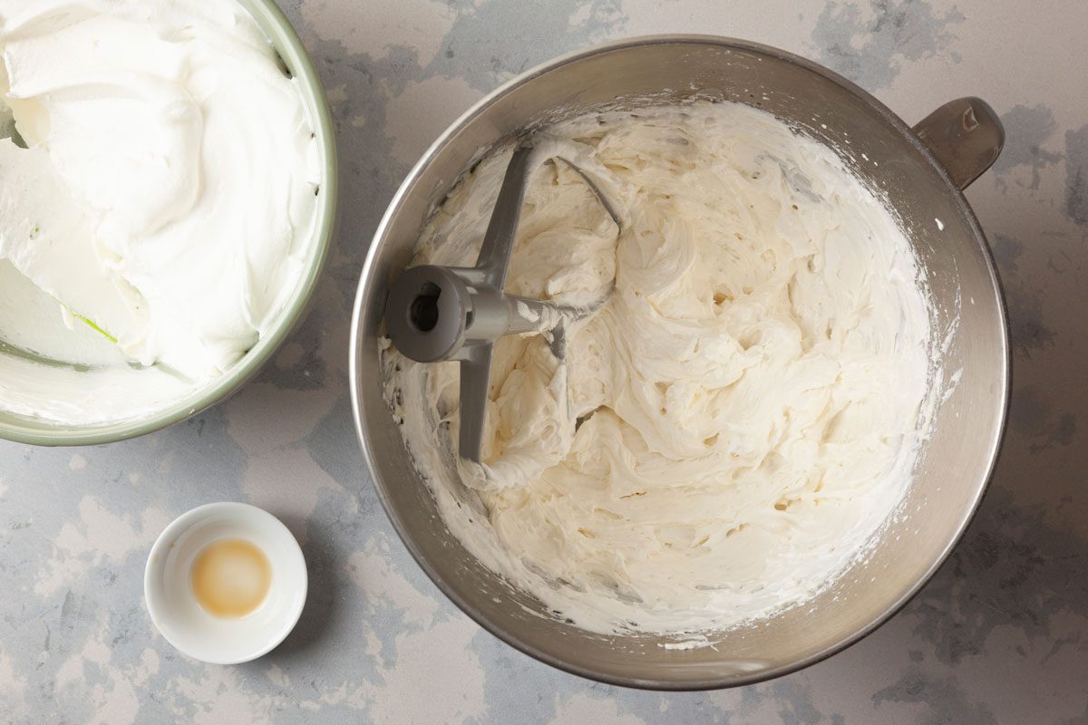 overhead shot of a steel mixing bowl filled frosting or whipped cream, on the left, a light green bowl contains white whipped cream, adjacent to these bowls is a small white dish that holds a liquid, possibly vanilla extract, set against a lightly gray surface