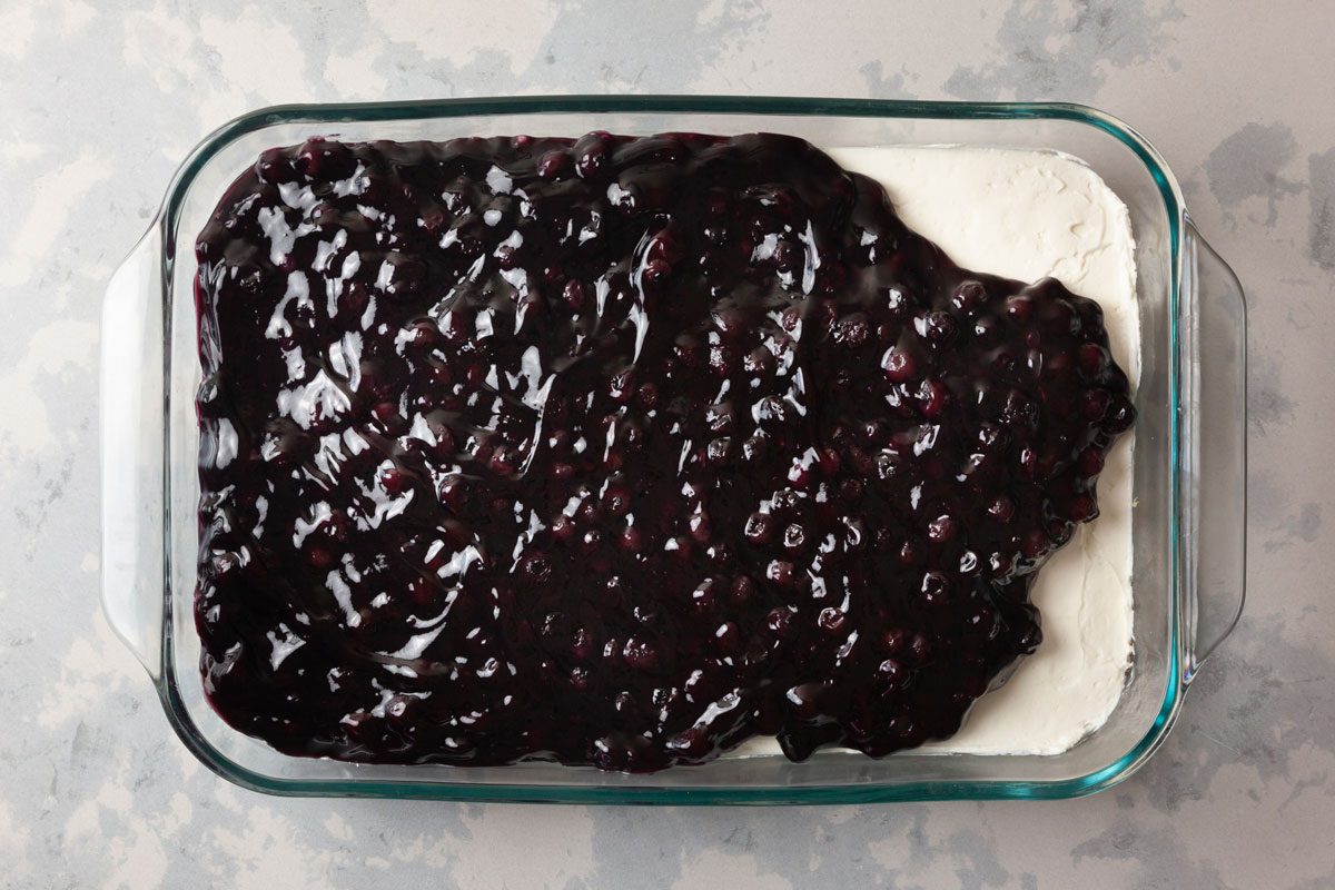 overhead shot of a glass baking dish filled with a dessert that has a creamy white layer at the bottom, topped with blueberry sauce; the dish sits on a light colored surface