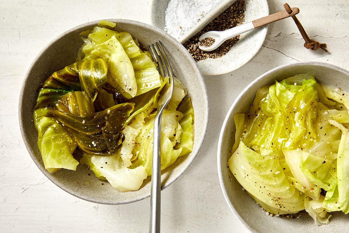 Two bowls of cooked cabbage with forks, seasoned with black pepper. A small bowl containing salt and a wooden spoon is placed nearby on a light surface.