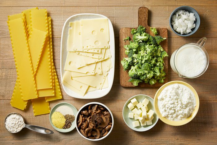 Ingredients for a lasagna: uncooked lasagna sheets, Swiss cheese slices, chopped broccoli, diced onion, a jug of milk, flour, grated cheese, sautéed mushrooms, butter cubes, and ricotta cheese in bowls on a table.