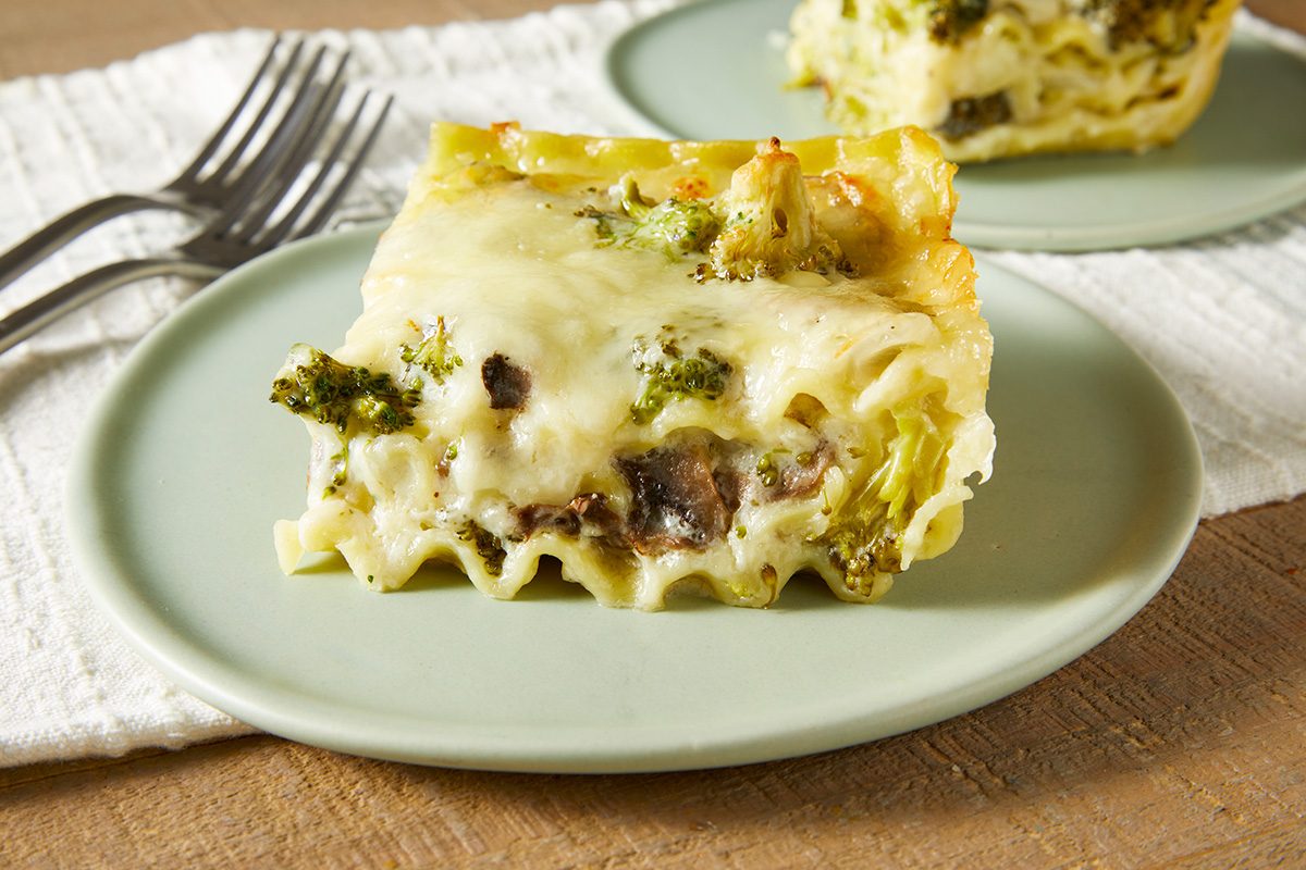 A slice of vegetable lasagna with visible layers of pasta, cheese, broccoli, and mushrooms sits on a light green plate. Another plate with the same dish is blurred in the background. Two forks are placed nearby on a white cloth.