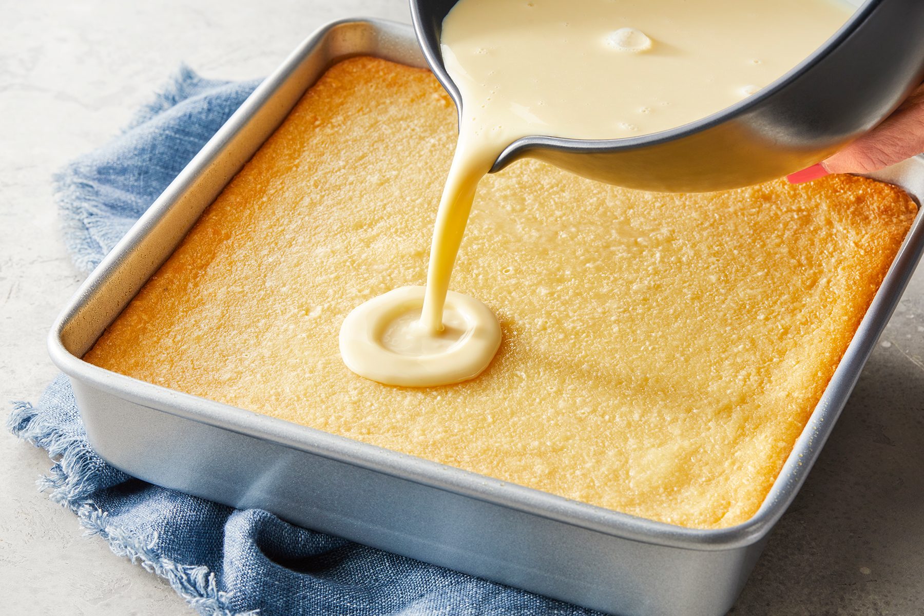 Batter is being poured from a bowl onto a square, baked yellow cake in a gray metal pan resting on a blue cloth. The cake has a smooth, golden surface.