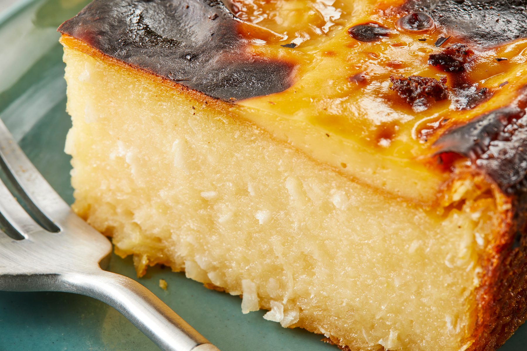 Close-up of a slice of custard pie with a golden-brown top and creamy filling, served on a plate. A fork is placed beside it, ready for use. The pie's surface shows light caramelization.