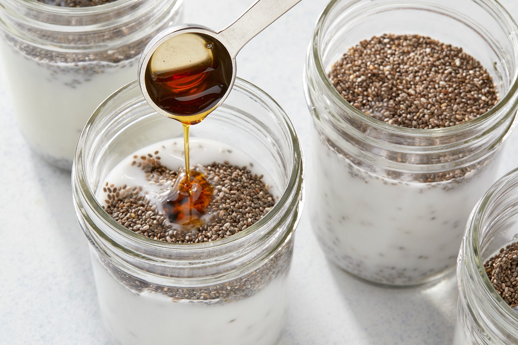 A spoon drizzles honey into a glass jar filled with yogurt and chia seeds. Several similar jars with chia seeds and yogurt are present in the background, all placed on a light-colored surface.