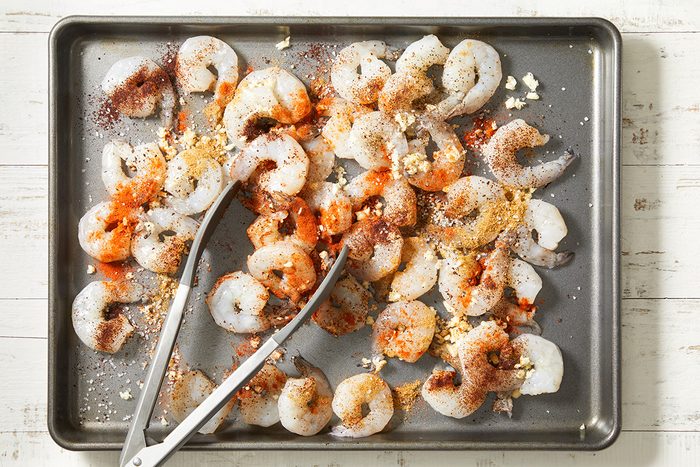 Raw shrimp on a metal baking sheet, sprinkled with various spices and minced garlic, with a pair of metal tongs resting among the shrimp.