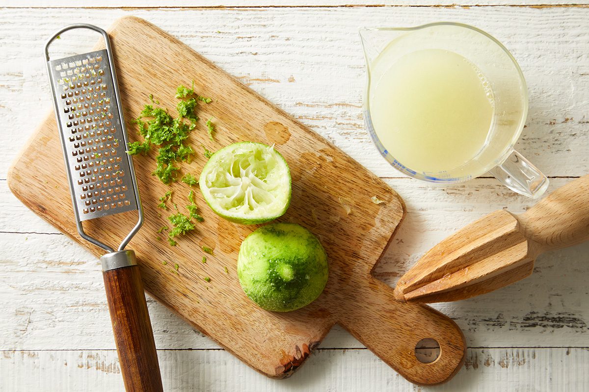 A wooden cutting board holds a zester with lime zest, a halved squeezed lime, and a wooden citrus reamer. Next to the board is a glass measuring cup filled with lime juice on a white wooden surface.