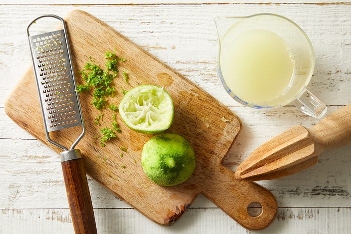 A wooden cutting board holds a zester with lime zest, a halved squeezed lime, and a wooden citrus reamer. Next to the board is a glass measuring cup filled with lime juice on a white wooden surface.