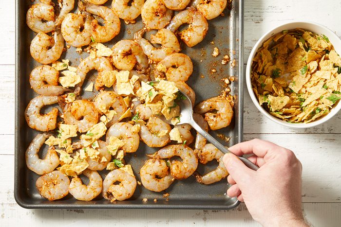 A hand sprinkles crushed tortilla chips over raw seasoned shrimp arranged on a baking sheet, with a bowl of crushed chips nearby on a white wooden surface.
