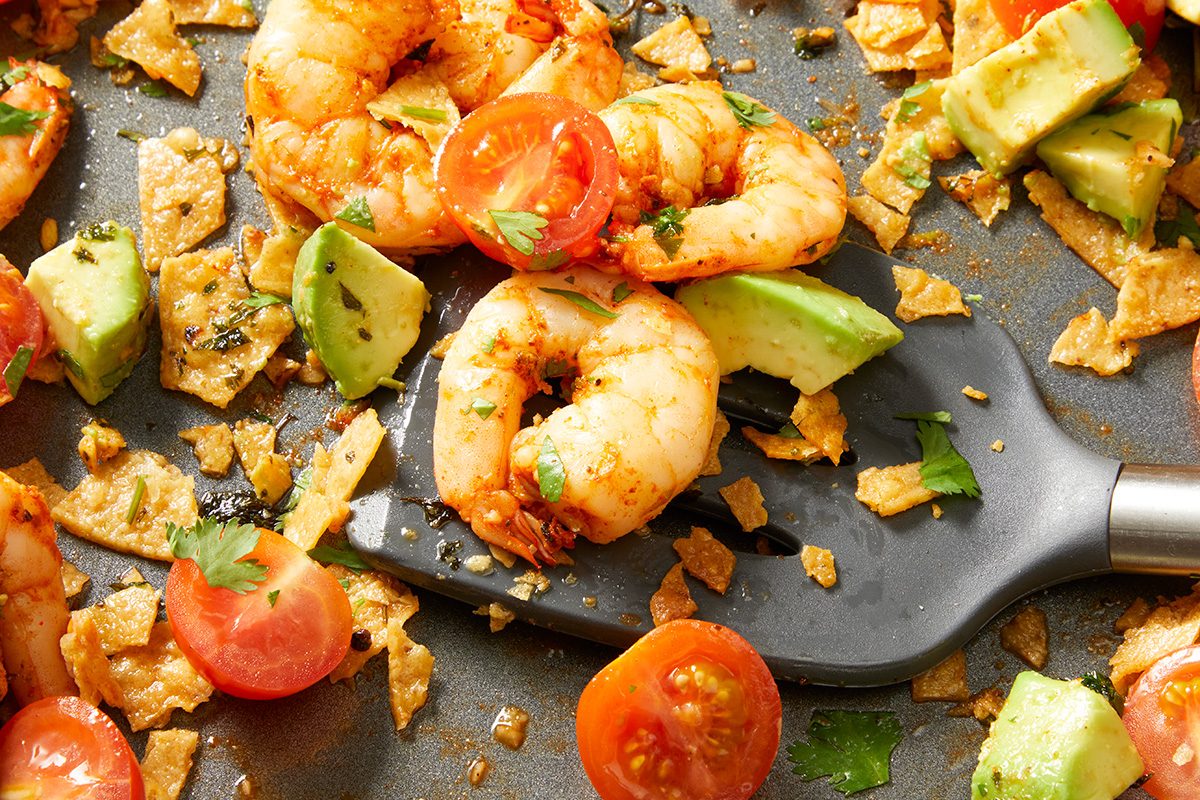 A spatula lifts seasoned shrimp, cherry tomato halves, avocado chunks, and crispy tortilla pieces from a pan, garnished with fresh herbs.