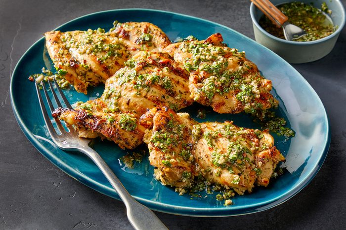 shot of Chimichurri Chicken, plated on a turquoise oval dish; a fork rests beside the chicken, in the background, a small bowl holds additional chimichurri sauce