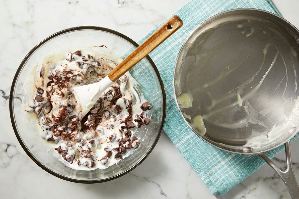 A glass bowl on a marble surface contains a mixture of chocolate chips and cream, being stirred with a spatula.