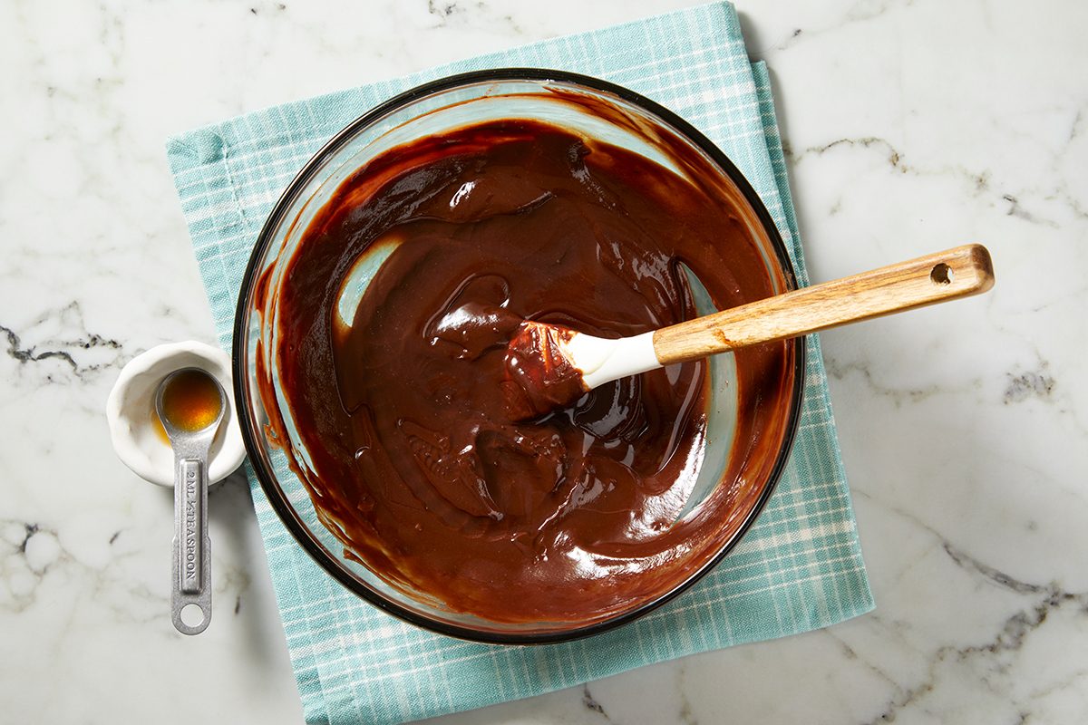 A glass bowl filled with smooth, melted chocolate with a wooden spatula rests on a teal checkered cloth.