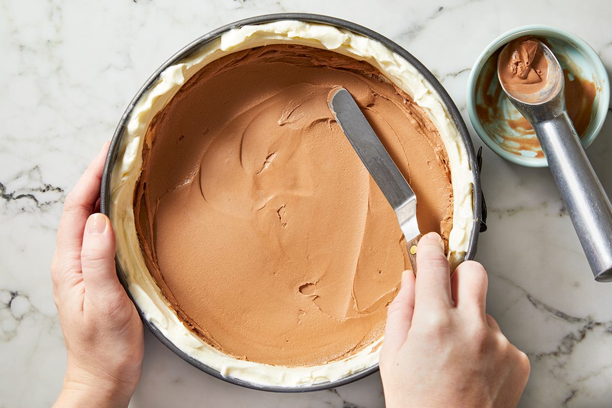 A person smooths chocolate ice cream in a round cake pan lined with white cream, using a spatula.
