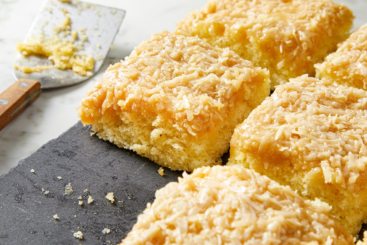Slices of coconut cake topped with shredded coconut are arranged on a dark slate surface. A spatula with crumbs rests nearby, all set on a white countertop.