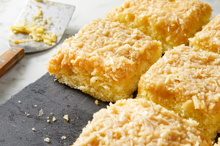 Slices of coconut cake topped with shredded coconut are arranged on a dark slate surface. A spatula with crumbs rests nearby, all set on a white countertop.
