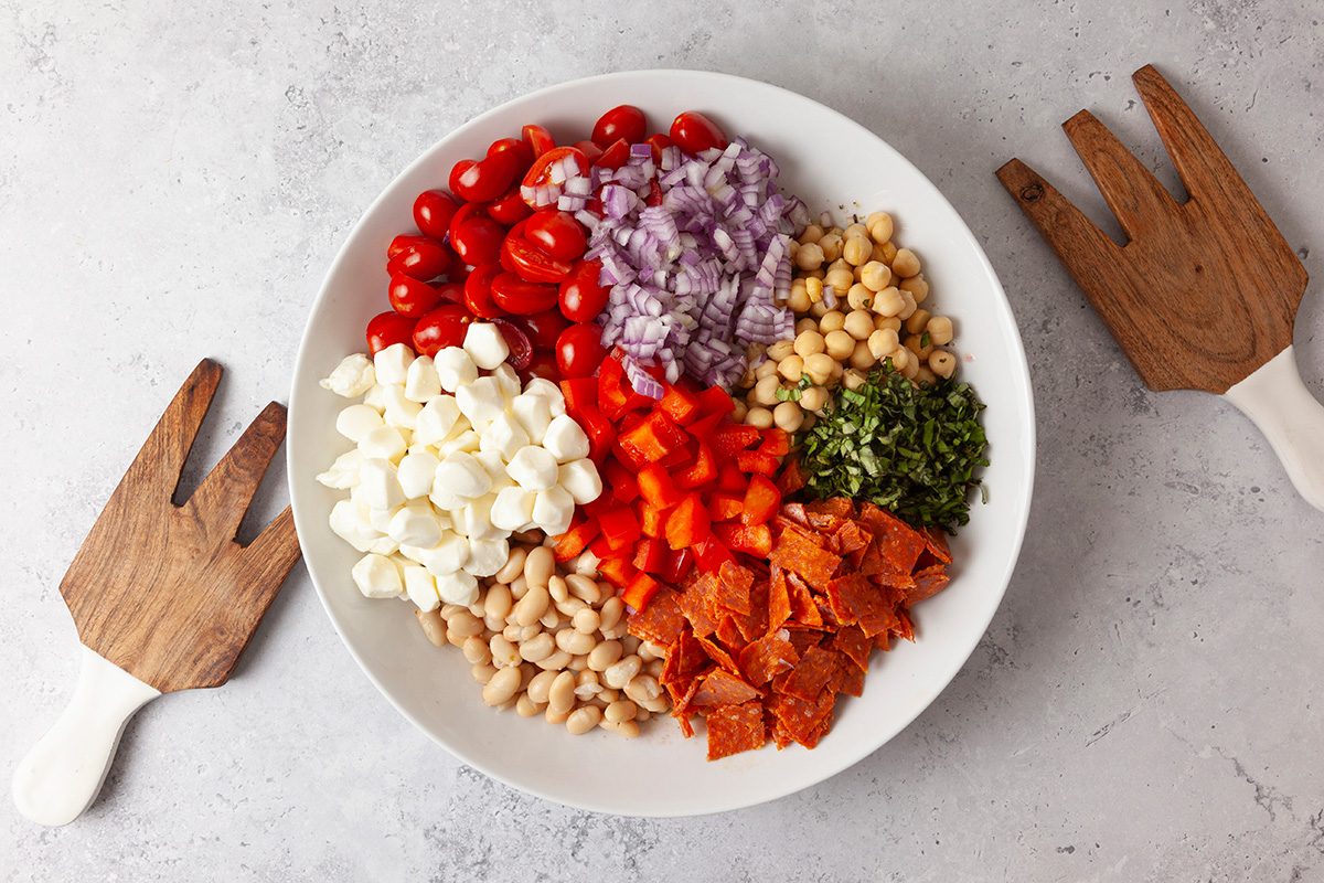 A white bowl filled with chopped red onions, grape tomatoes, chickpeas, mozzarella balls, red bell pepper, white beans, pepperoni slices, and chopped herbs, placed on a light surface with wooden salad servers beside it.