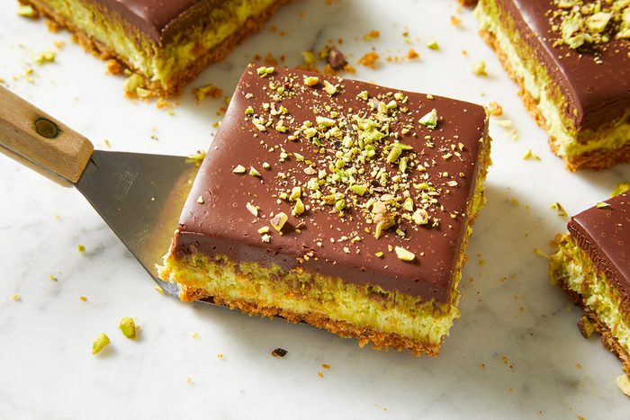 A spatula lifts a square dessert bar with a chocolate topping, chopped pistachios, and layered cookie and creamy filling on a marble surface. Other dessert bars are visible in the background.