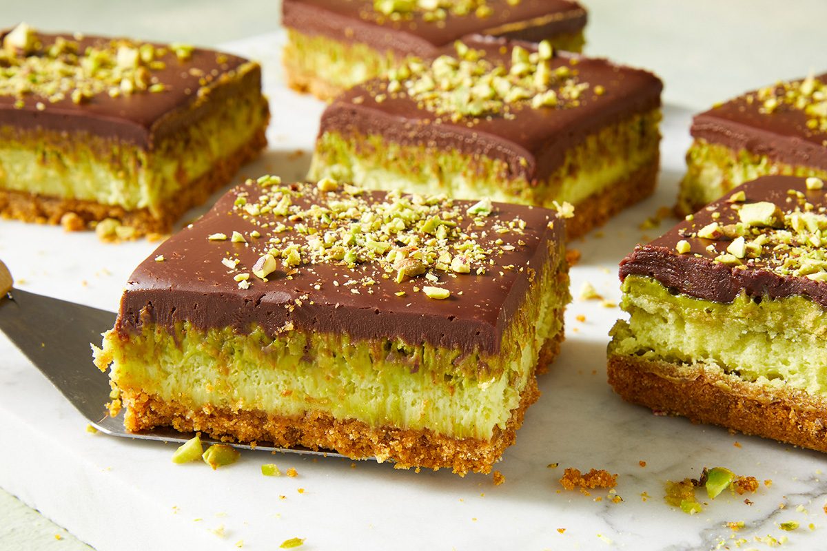 Six square dessert bars with a crumbly crust, creamy green pistachio layer, and a thick layer of chocolate on top, garnished with chopped pistachios, are arranged on a marble surface.