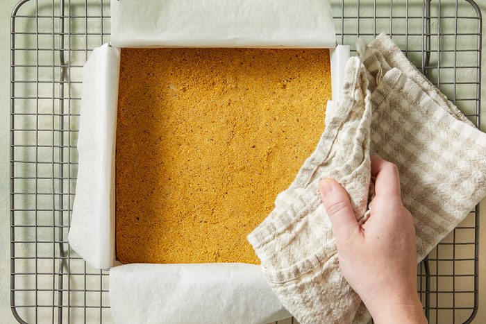 A hand using a checkered cloth lifts a square pan lined with parchment paper, filled with a golden-brown baked cake, resting on a wire cooling rack.