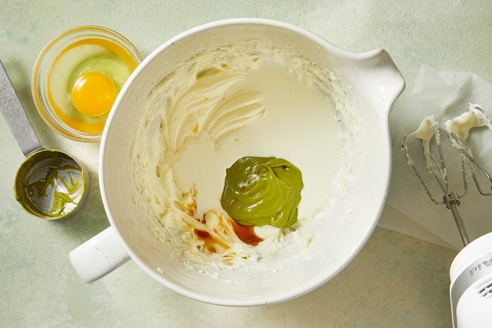 A mixing bowl contains creamed ingredients, matcha powder, and vanilla extract. An egg in a small bowl, a metal spoon, and a mixer with a whisk attachment are nearby on a light-colored surface.