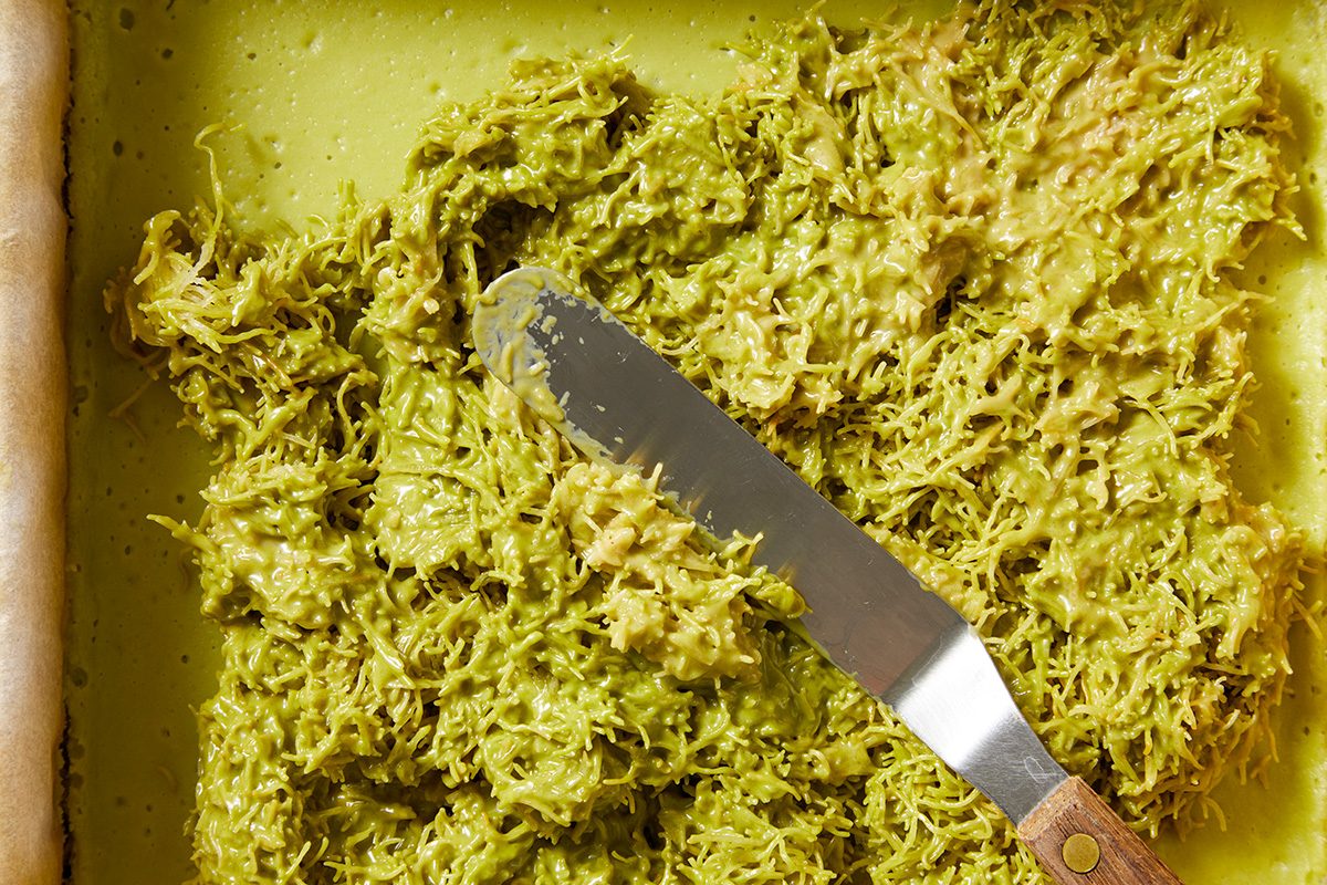 A close-up of shredded green filling being spread with a metal spatula on a rectangular crust, possibly for a dessert or savory dish. The filling appears textured and slightly creamy.