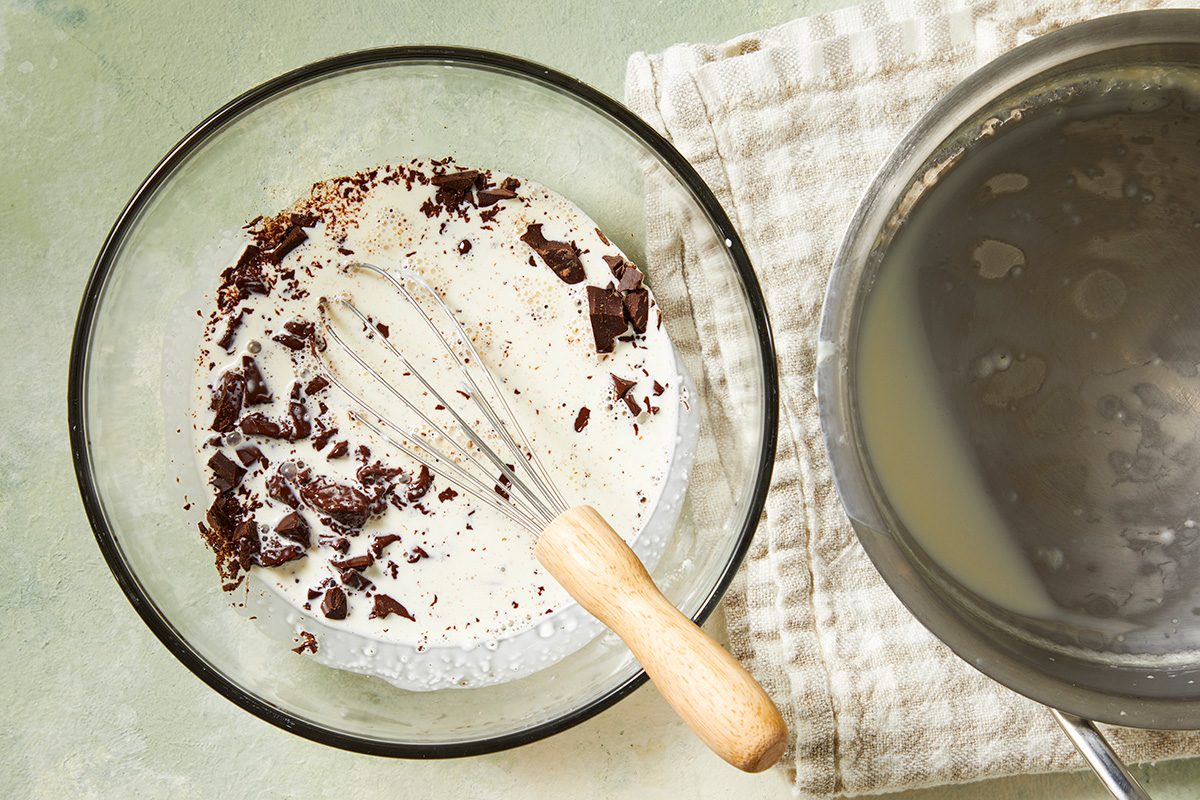 A glass bowl with cream and chopped chocolate being whisked, set next to a saucepan with liquid and a folded checkered dish towel on a green surface.