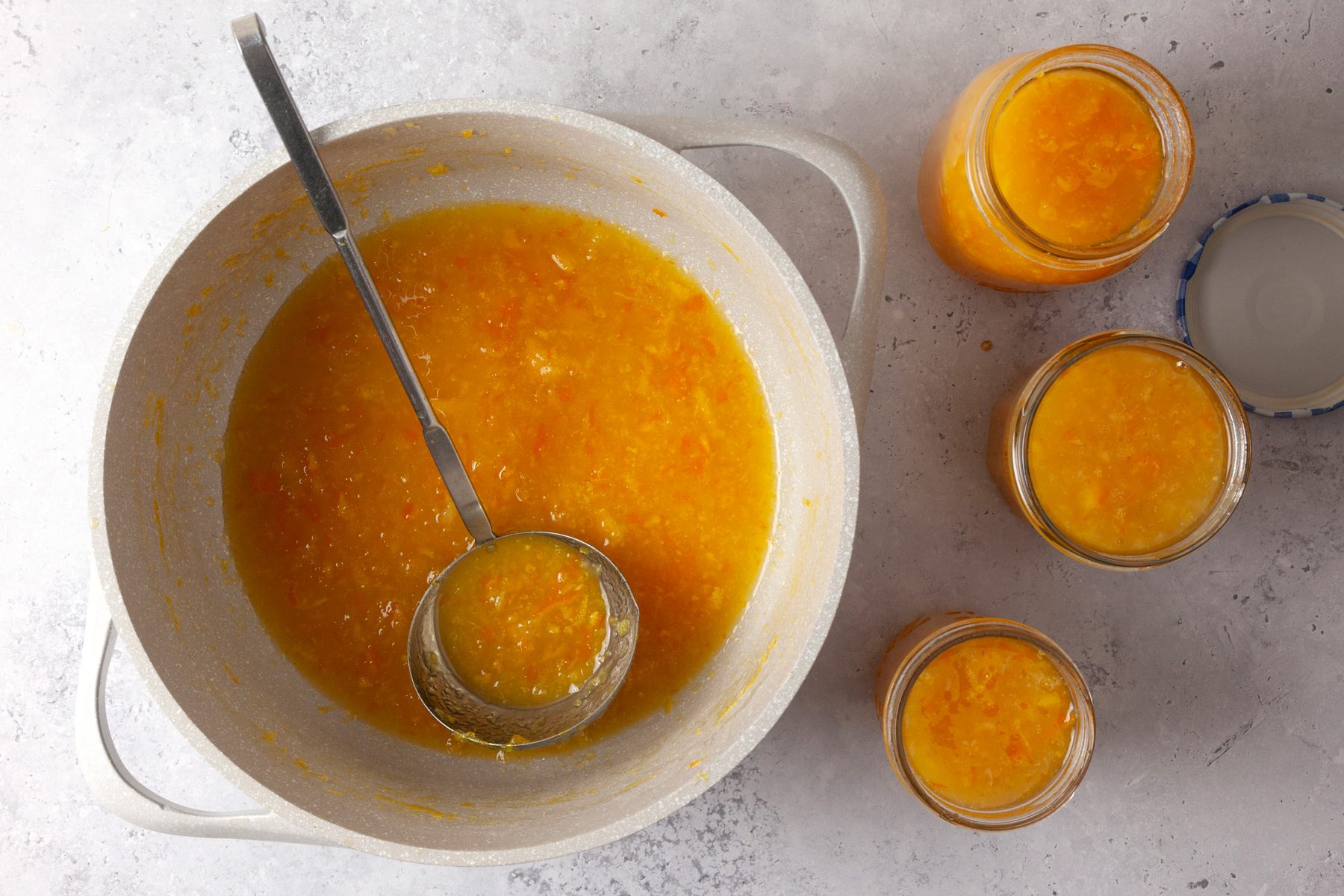 overhead shot of a large pot filled with an orange mixture; a ladle rests inside the pot, surrounding the pot are several glass jars filled with the same orange mixture, the background has a subtle gray surface