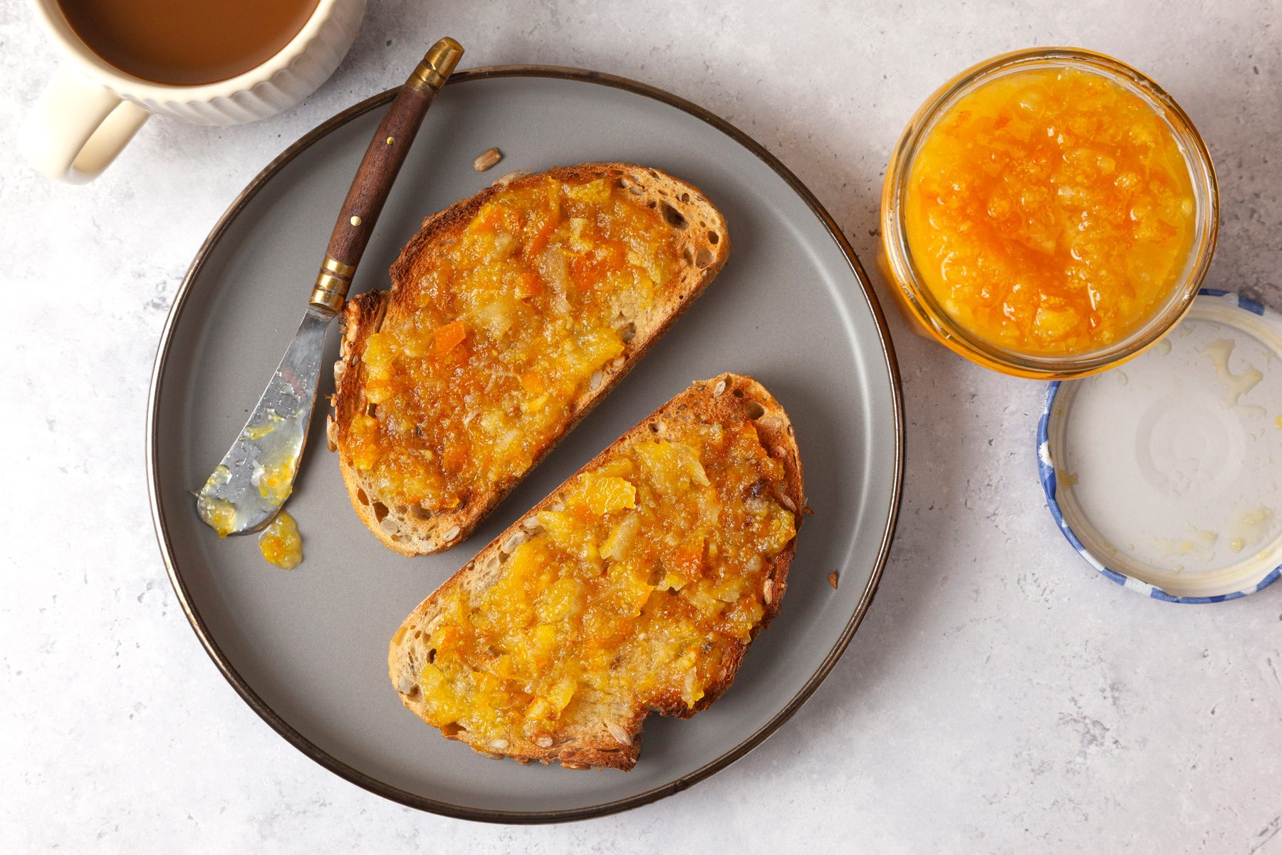 overhead shot of two slices of toast topped with a spread of orange marmalade on a round grey plate; A small butter knife with a wooden handle rests beside the toast; In the background, there is a cup of coffee sitting on a light colored surface