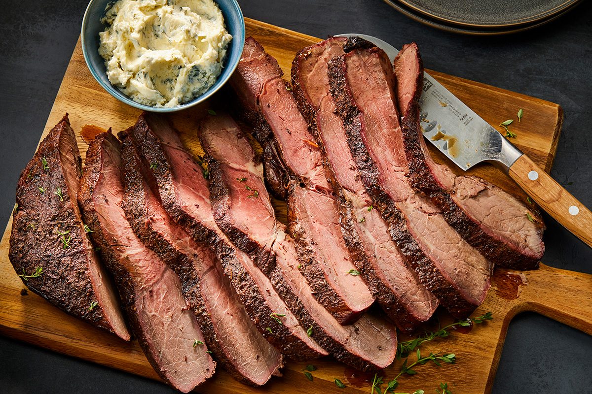 Horizontal AP; Pull shot of Grilled Sirloin Tip Roast, sliced into thin, even portions, the sliced roast is arranged on a wooden cutting board with a cutting knife placed beside, garnished with fresh herbs; beside the meat, there is a small bowl containing creamy mashed potatoes