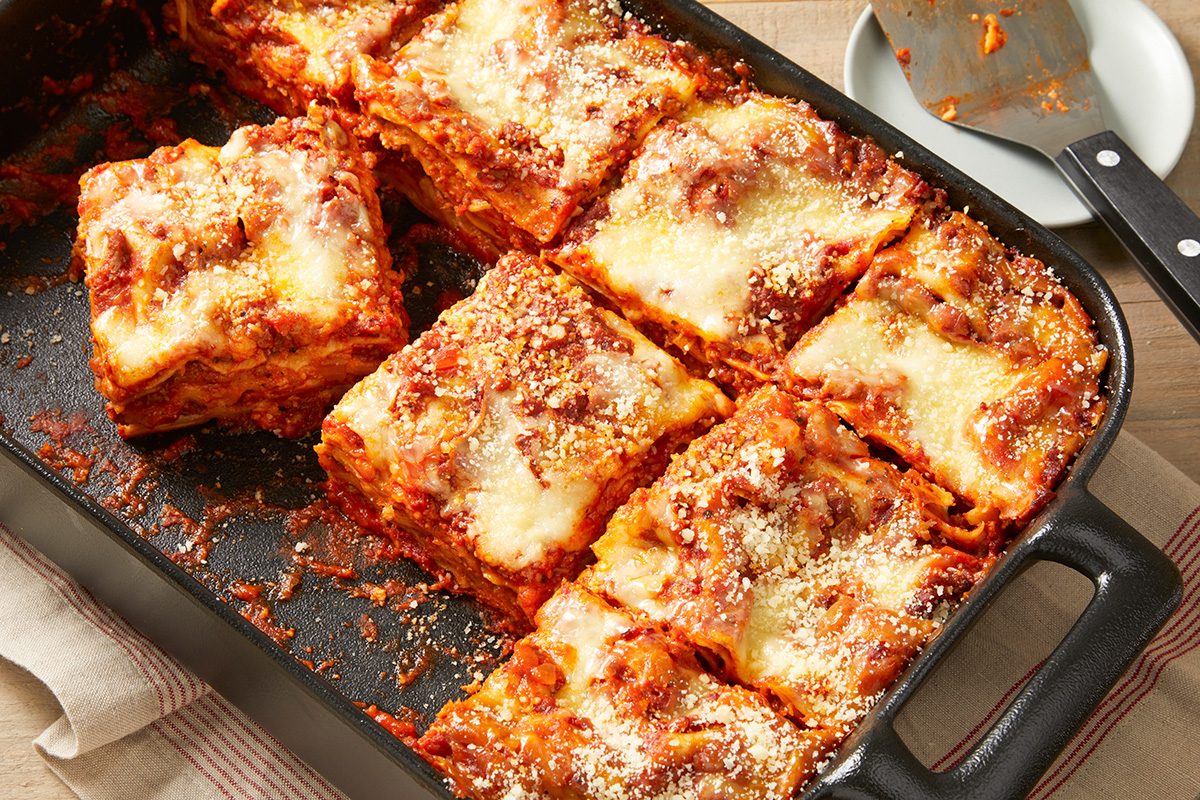 A rectangular baking dish filled with sliced lasagna. The lasagna has layers of pasta, tomato sauce, and melted cheese on top. A serving spatula rests nearby, and a piece of lasagna is slightly lifted.