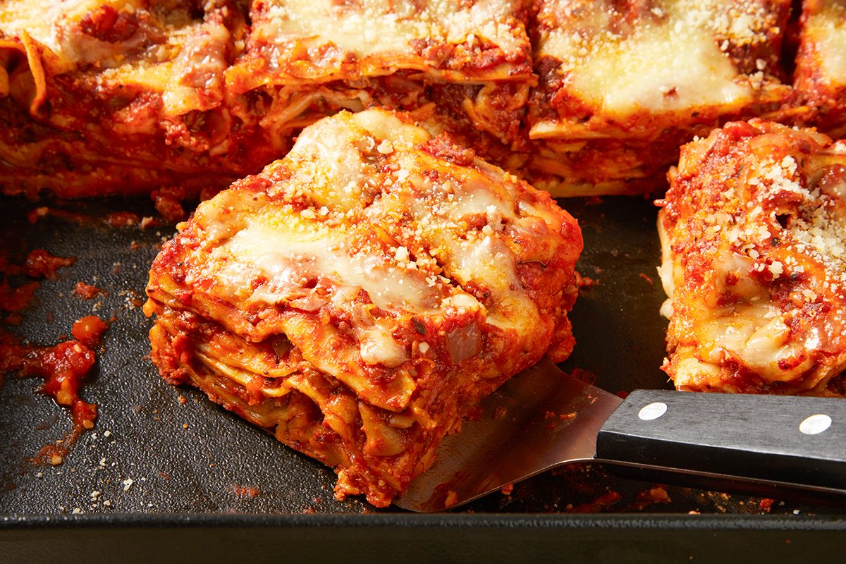 A close-up of a serving of lasagna on a spatula resting in a baking dish. The lasagna has layers of pasta, rich tomato sauce with meat, and melted cheese, topped with grated parmesan. Some slices remain in the dish.