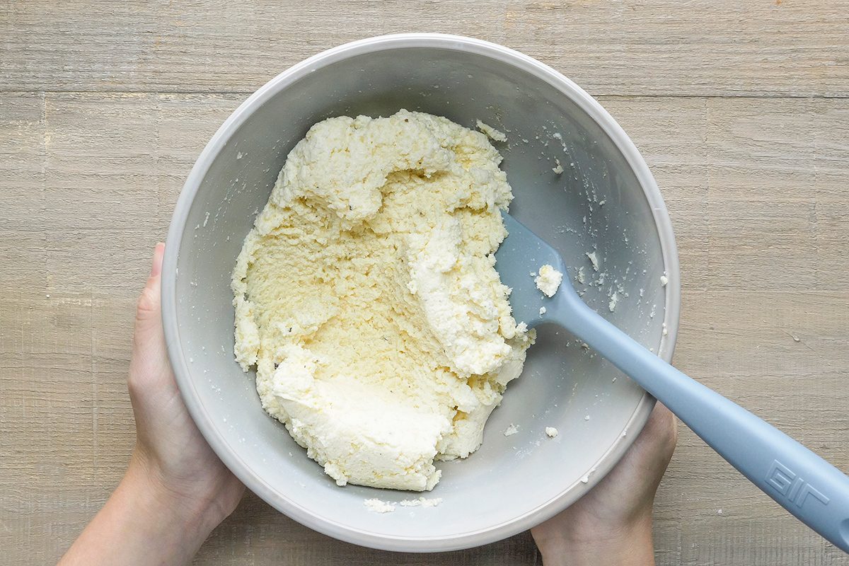 Hands holding a bowl with a mixture of creamy batter being stirred with a blue spatula. The bowl is placed on a wooden surface.