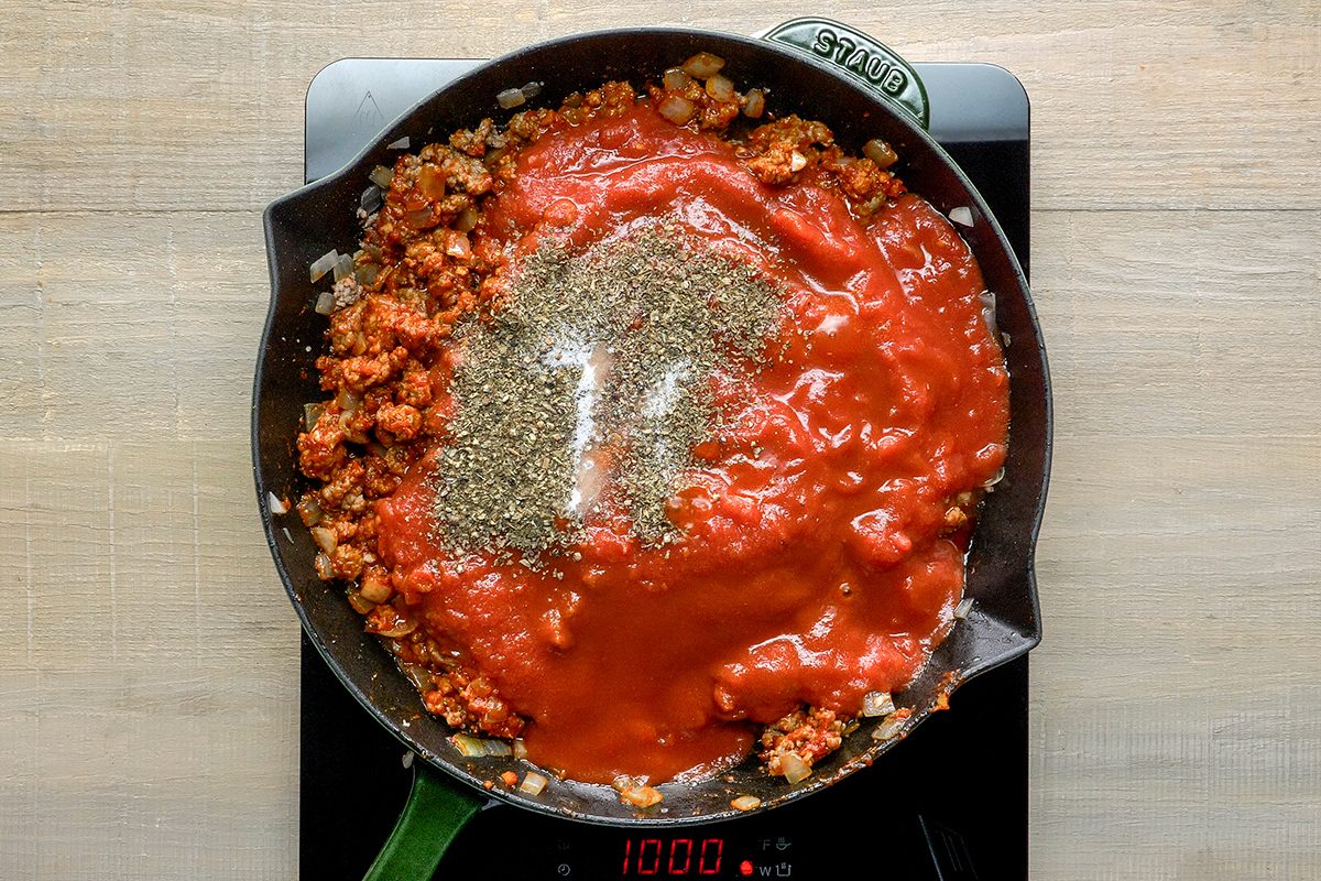 A skillet on an electric stove contains a tomato sauce mixture with browned meat and herbs, possibly Italian seasoning, sprinkled on top. The stovetop display reads 10:00. The skillet handle is green.