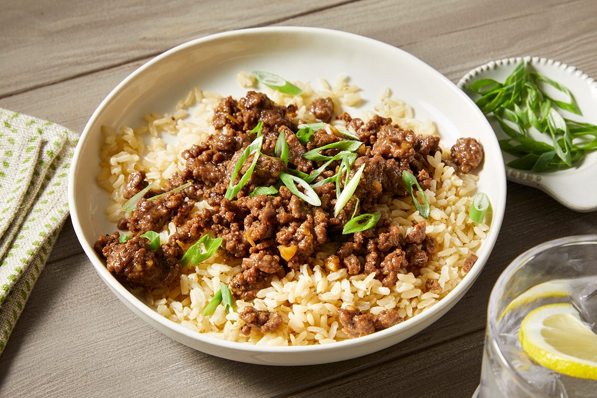 A white bowl filled with cooked rice topped with browned ground beef and garnished with sliced green onions. 