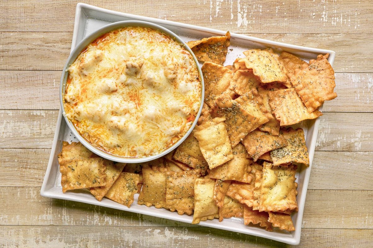 overhead shot of Lasagna Dip served in a circular bowl placed on a rectangular platter; the dip is topped with melted cheese, surrounding the dip are crispy, golden brown ravioli chips, the background is wooden