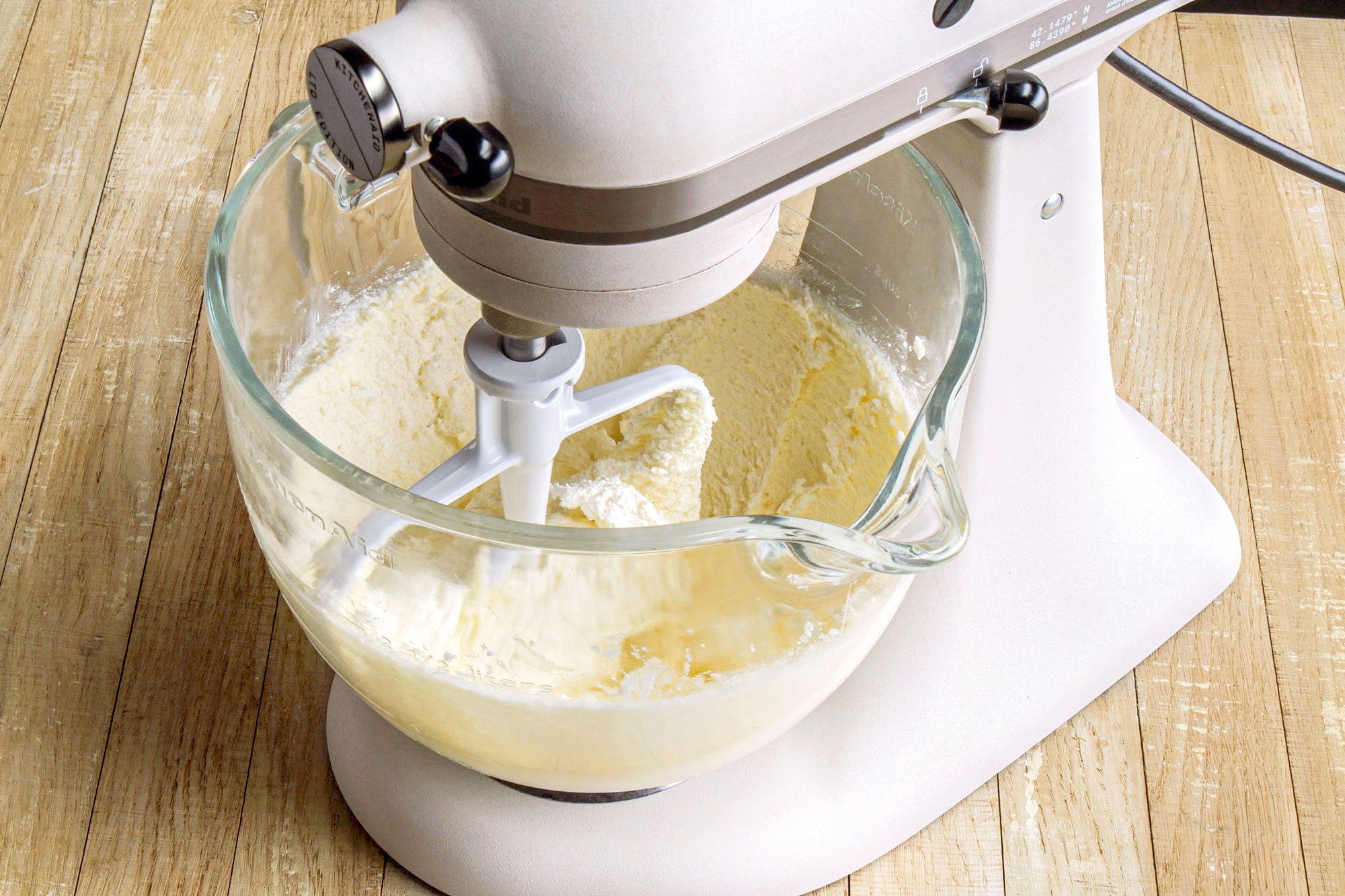 3/4th shot of a stand mixer positioned on a wooden surface; a glass mixing bowl filled with creamy batter or a similar mixture, the mixing attachments are visible inside the bowl, actively blending the ingredients; the surrounding wooden surface