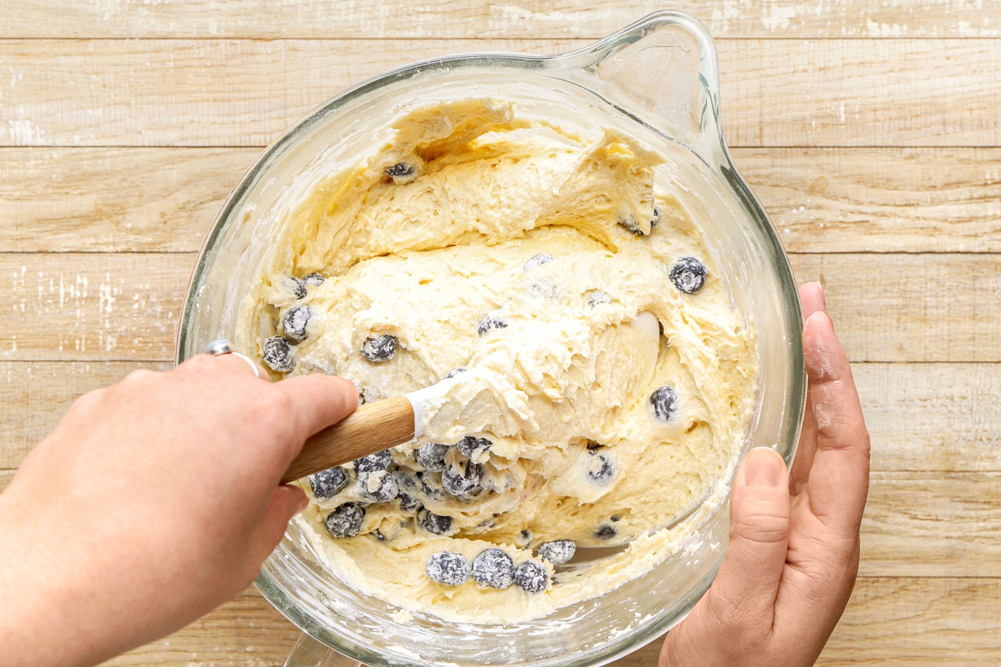 overhead shot of a glass mixing bowl filled with a creamy batter that has specks of blueberries scattered throughout; a wooden spoon is being used to mix the ingredients, with one hand grasping the spoon while the other hand supports the bowl; the background features a textured wooden surface